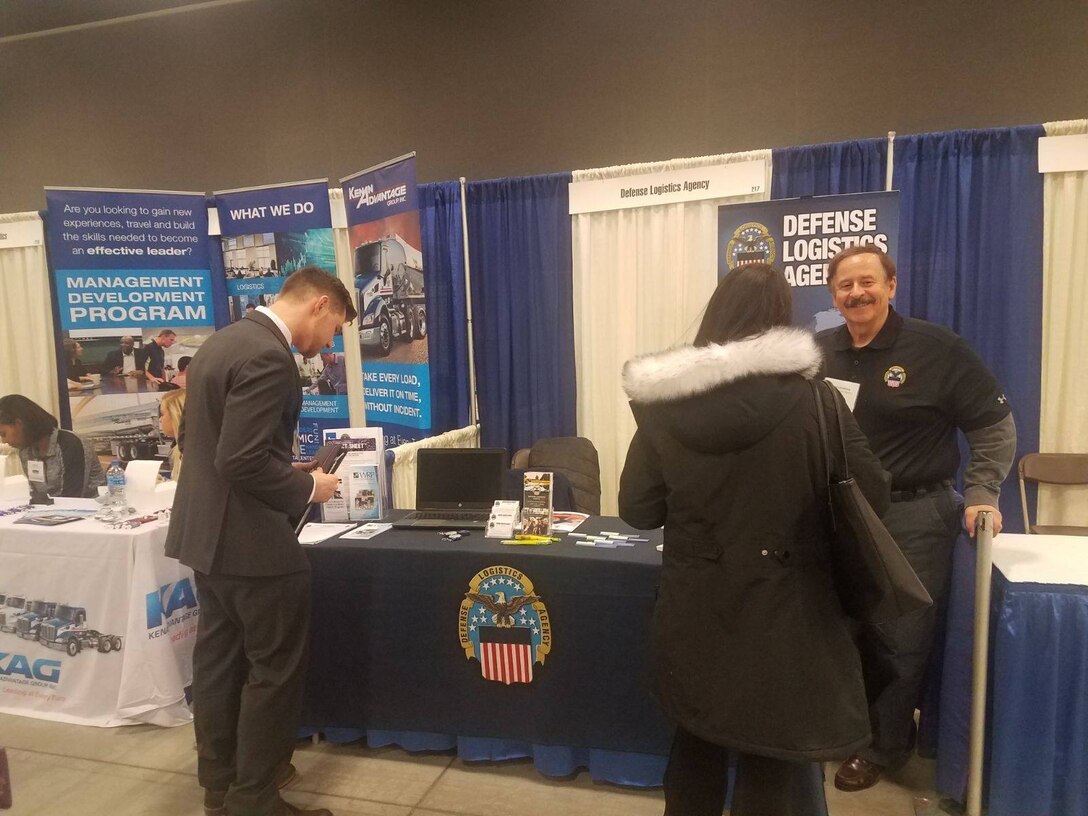 DLA employee stands behind an expo event table sharing information with a male and female college student