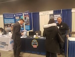 DLA employee stands behind an expo event table sharing information with a male and female college student