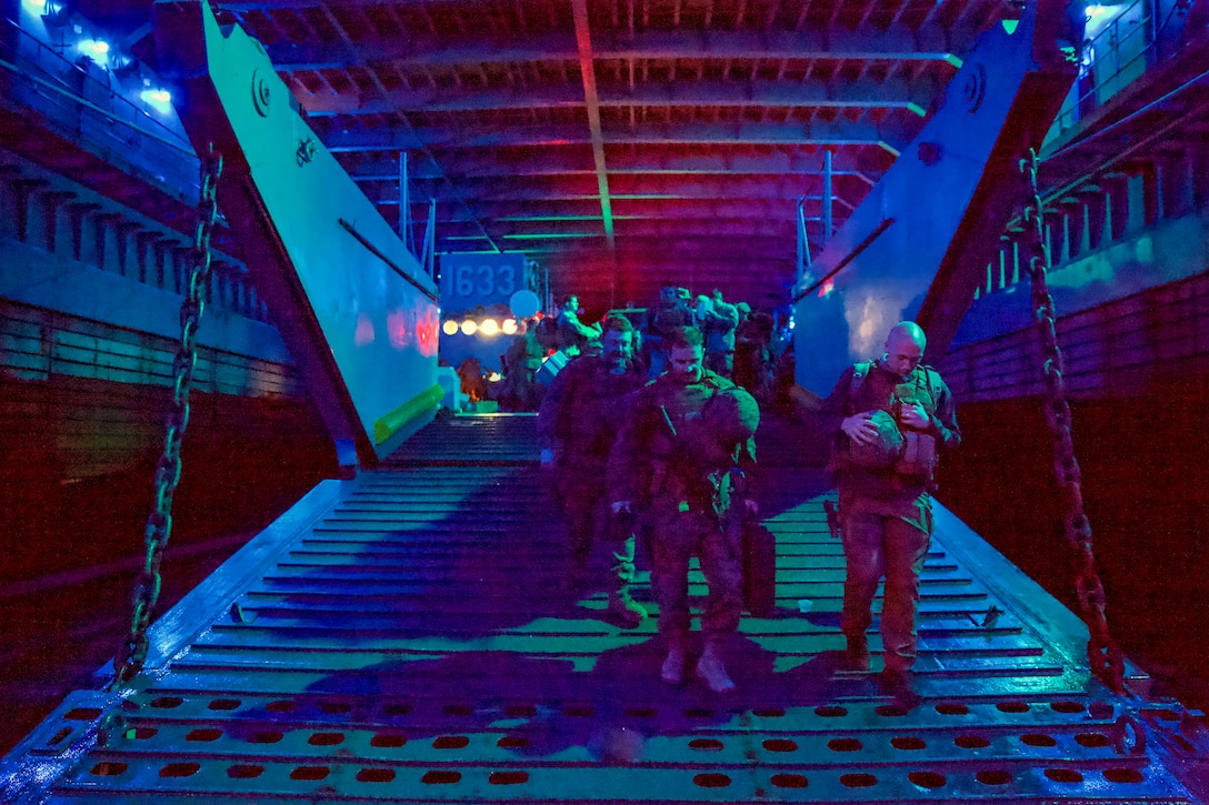 Marines and sailors walk out of the back of a landing ship at night.