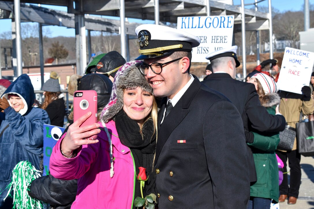 A sailor stands and smiles for a selfie with a smiling woman as they stand in a crowd.