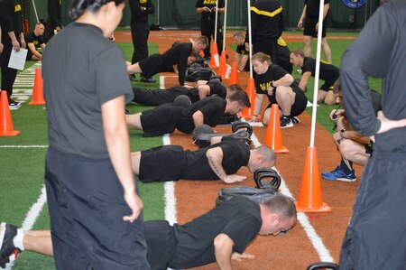 Soldiers in black shirts conducting physical training for the Army Combat Fitness Test. Soldiers conducting the Hand Release Push Up. Indoor stadium with orange and green turf