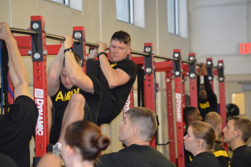 Soldiers in black shirts conducting physical training for the Army Combat Fitness Test. Pull Up bars are black with red beams with several Soldiers waiting in line for their turn.