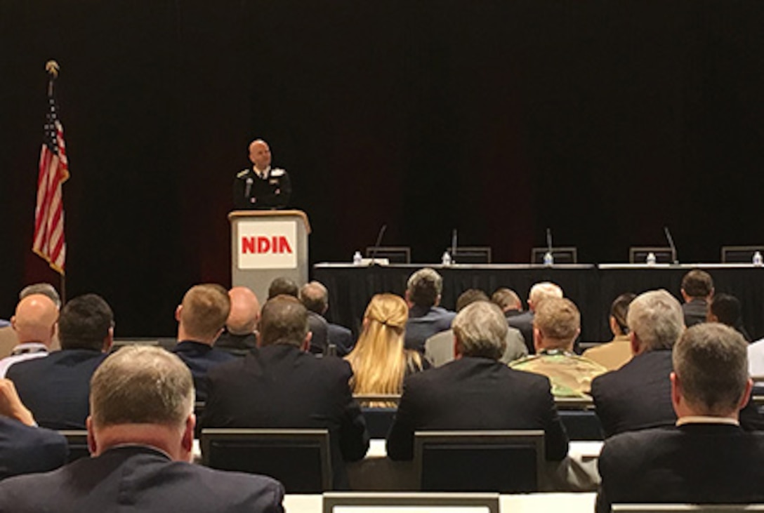 Navy Rear Adm. stands on stage behind a podium speaking to a crowded room