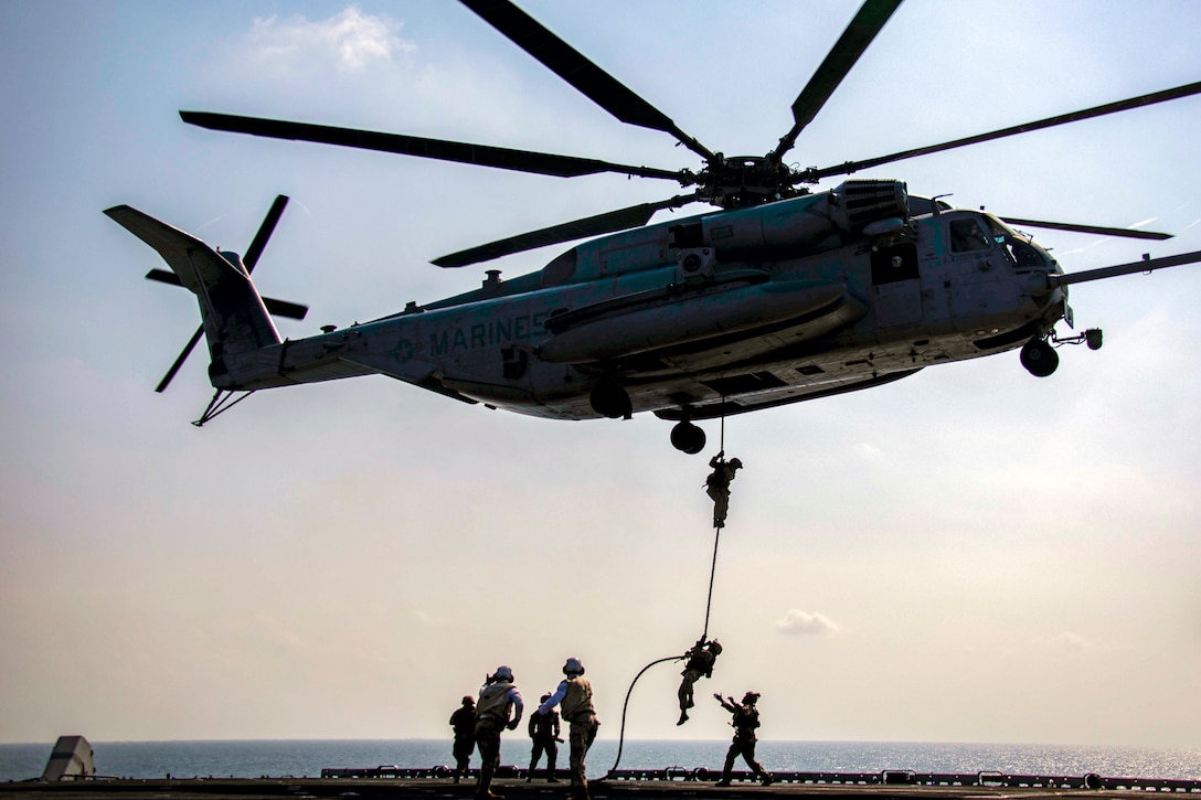 Marines climb down a rope hanging out of a helicopter onto the flight deck of a navy ship.