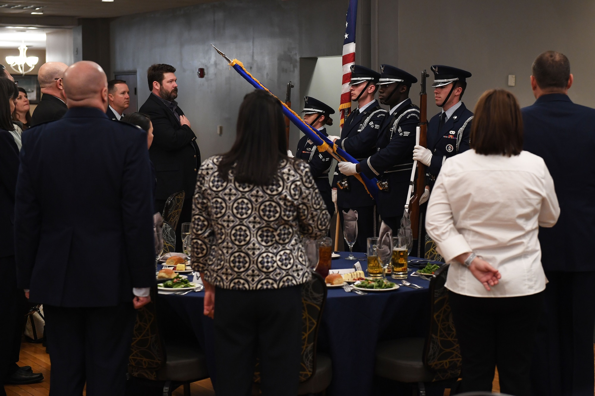 Men and Women stand, while honorguard members present the colors.