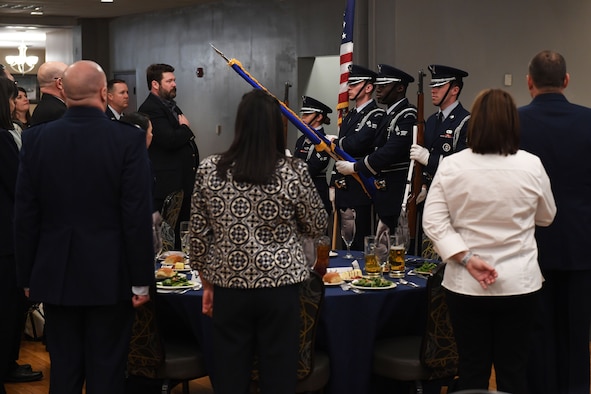 Men and Women stand, while honorguard members present the colors.
