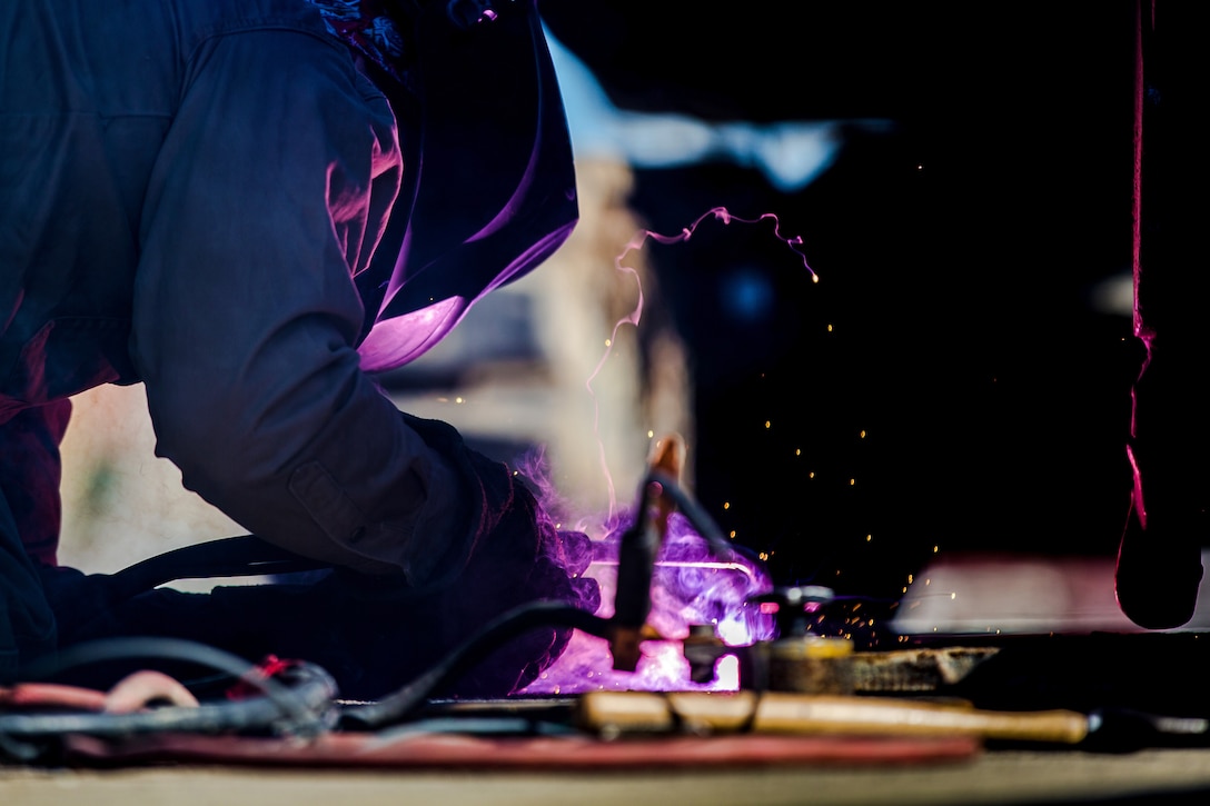 Sparks and purple smoke fly as a welder works on a steel plate.