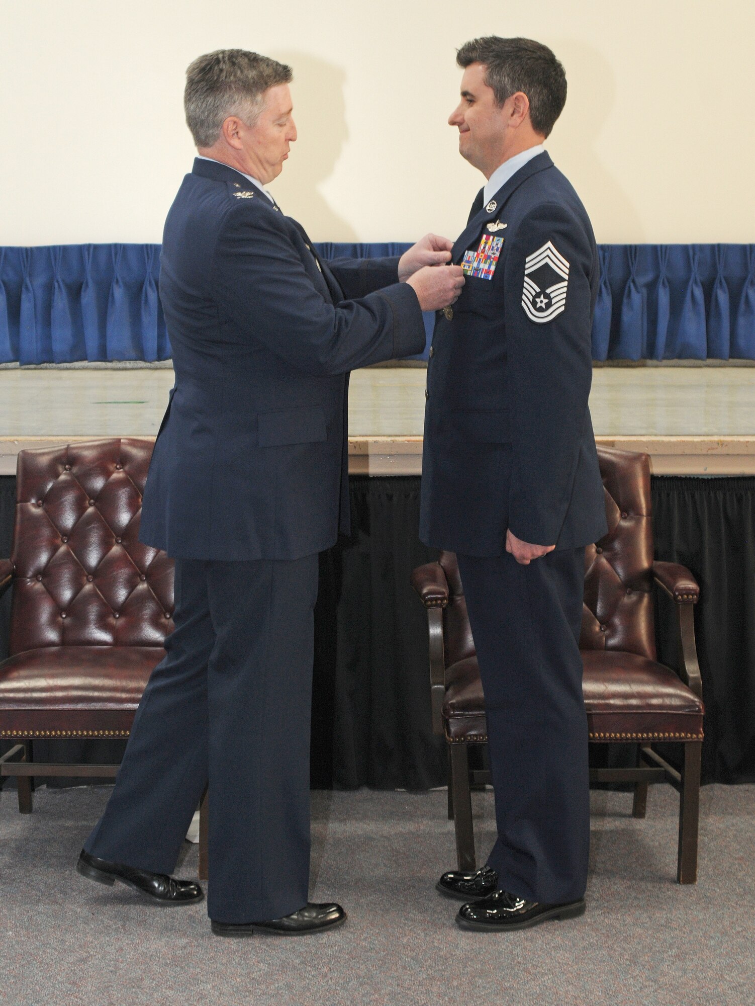 Two figures in profile, Col. reaching across to pin medal on Chief Master Sgt. against a curtain backdrop.
