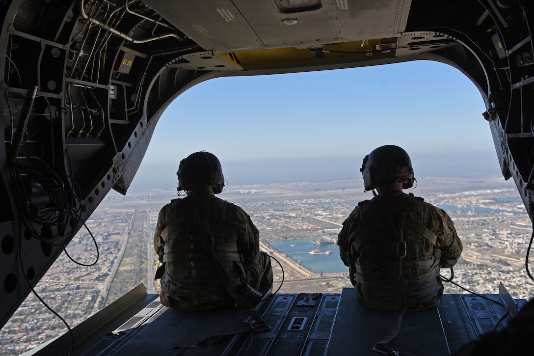 Two U.S. troops sit in a helicopter looking out over a city.