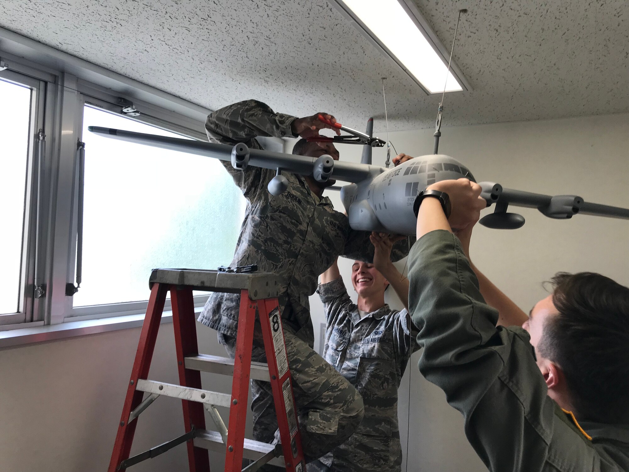 Master Sgt. Carlos Boglin, 374th OSS first sergeant (on ladder), Airman 1st Class Ricardo Bustamante, 374 OSS intelligence journeyman (background) and Capt. Duncan Dillon 374 OSS executive officer, hang a C-130J Super Hercules model in the OSS building at Yokota Air Base, Japan, Sept. 18, 2018.