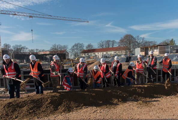 Group of adults and students in orange safety vests and hardhats shovel dirt in front of a construction site.