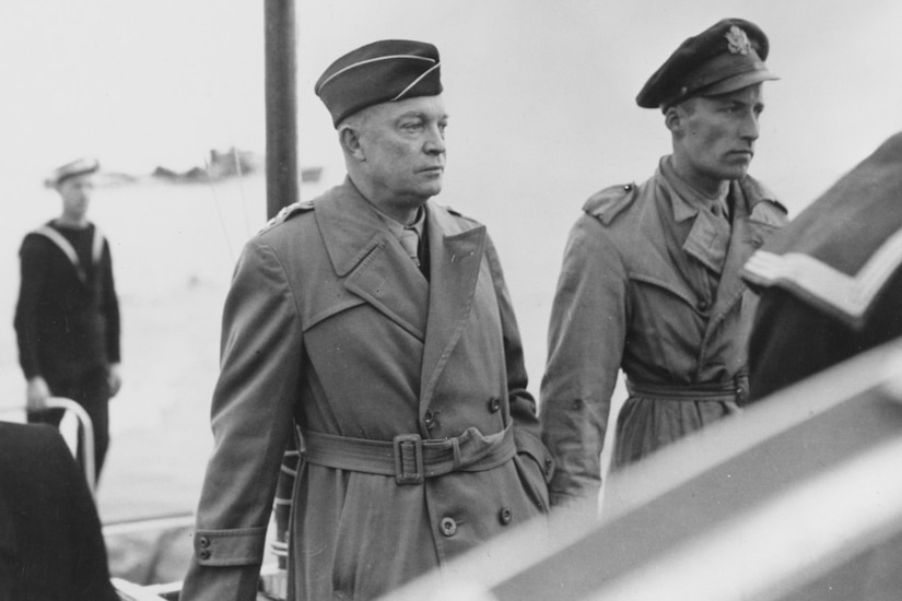 Two high-ranking officers stand on a boat with a sailor in the background.