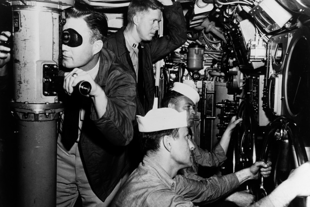 Two men stand and two men sit in a submarine control area.