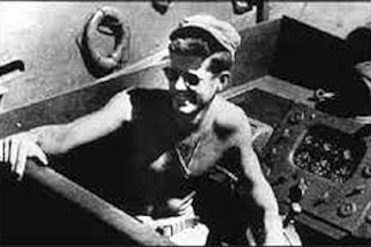 A shirtless John F. Kennedy smiles from the driver’s seat of a motor torpedo boat.