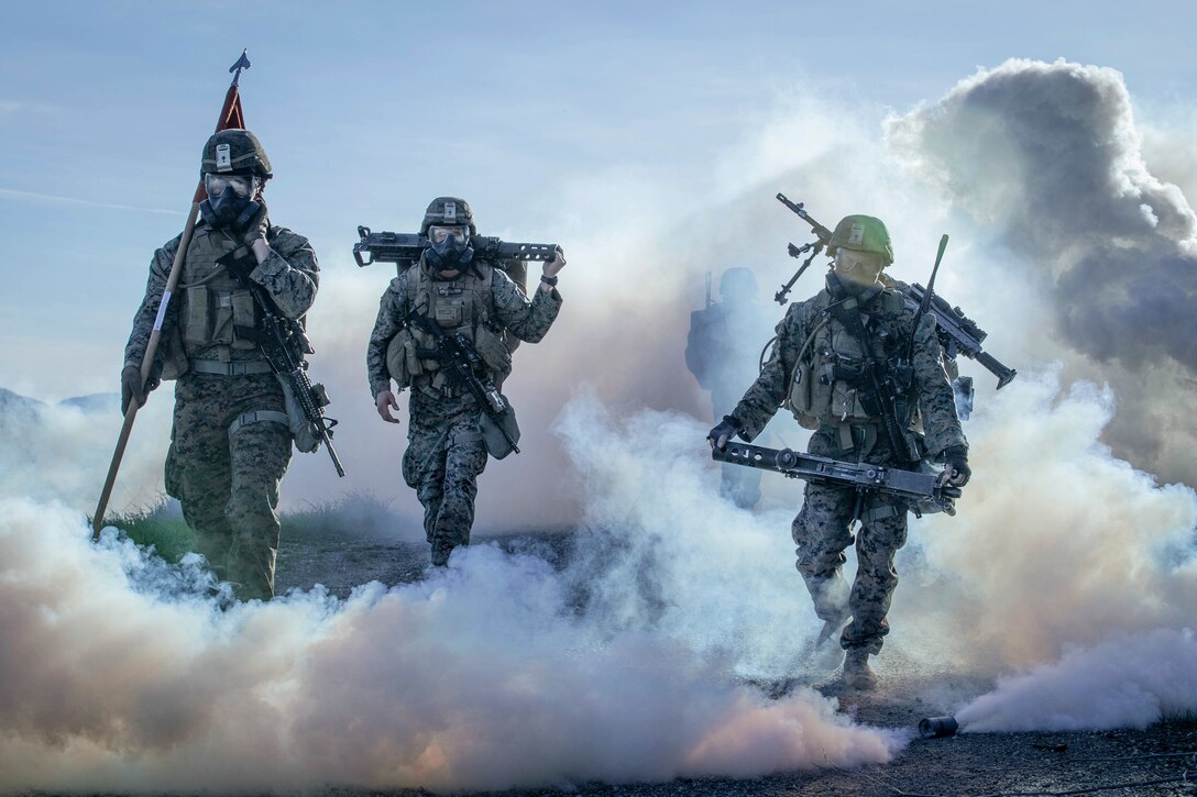 Marines in protective gear hike through smoke.
