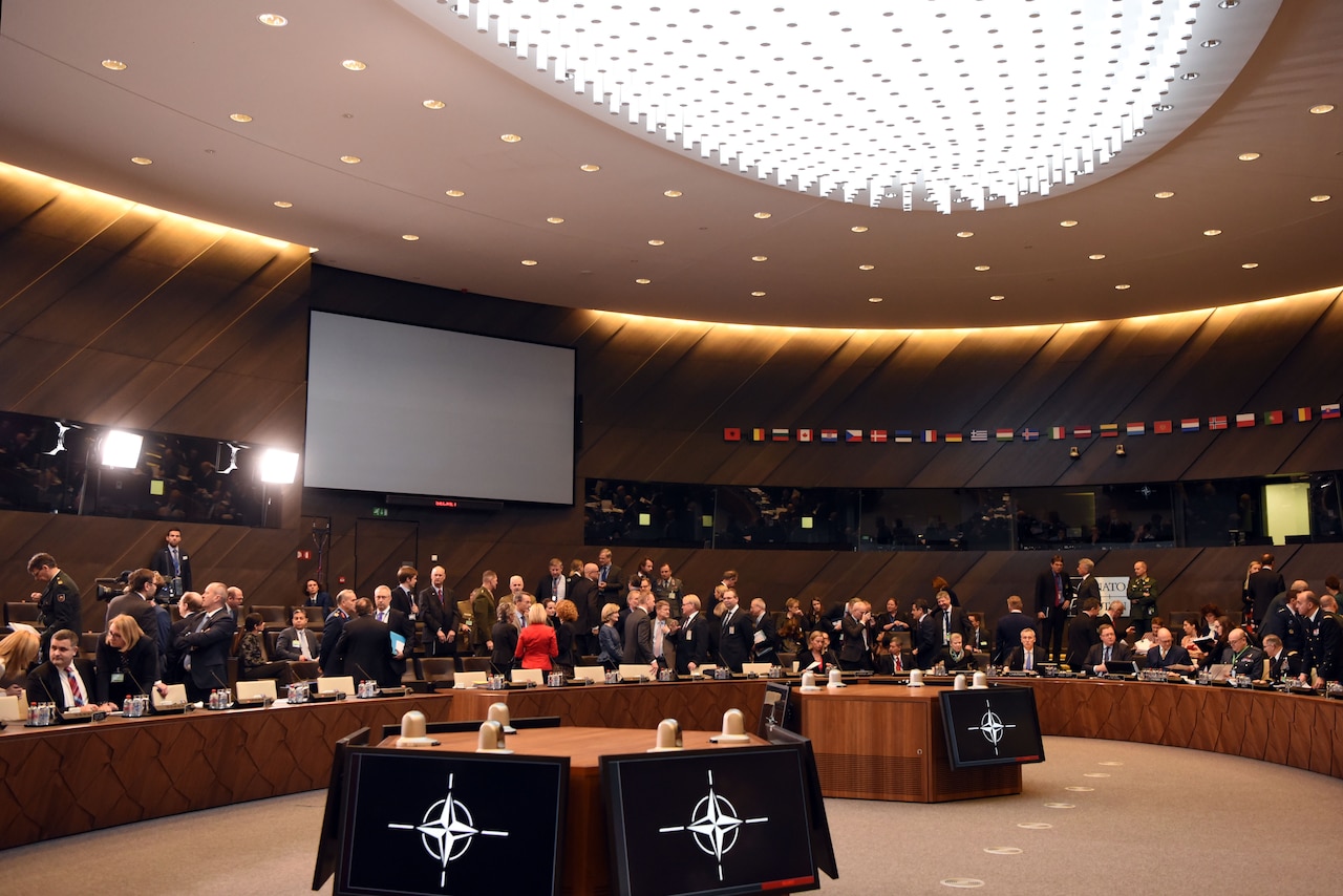 Defense ministers sit at curved tables in a large meeting room.