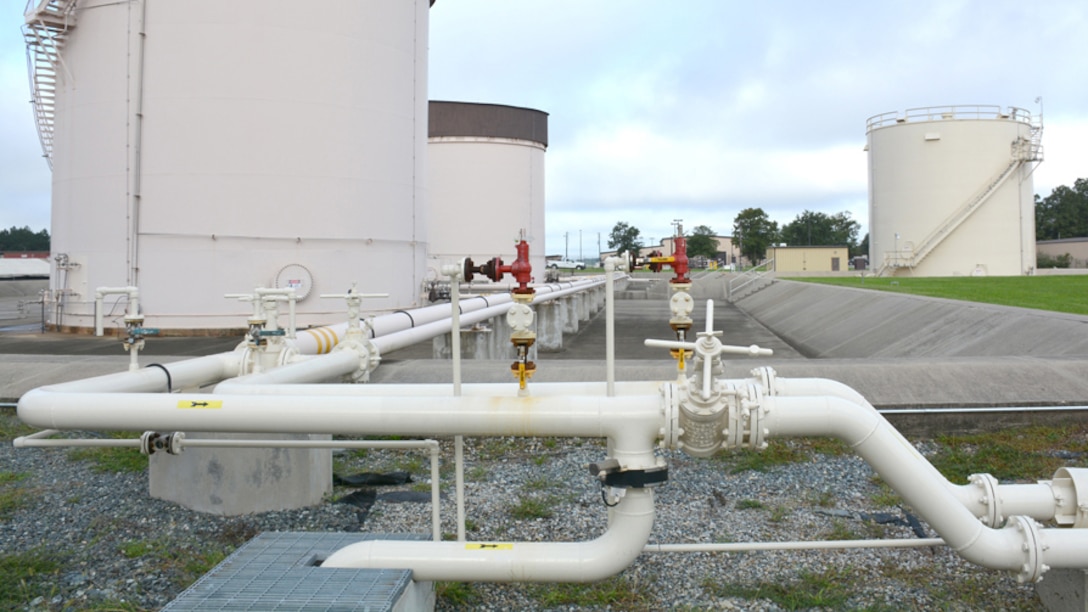 Three large, outdoor cylindrical fuel tanks in the background are connected with sets of pipes extending into the foreground.