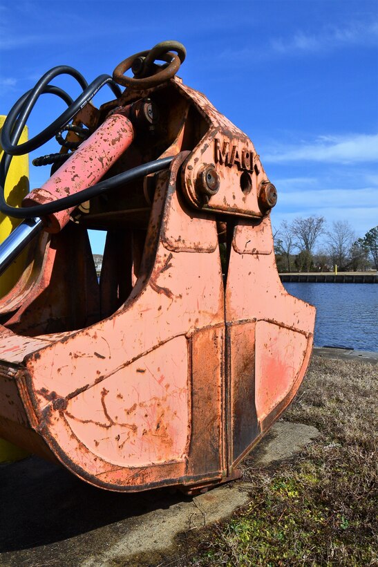 a red and weathers clamshell crane bucket sits along the waterway