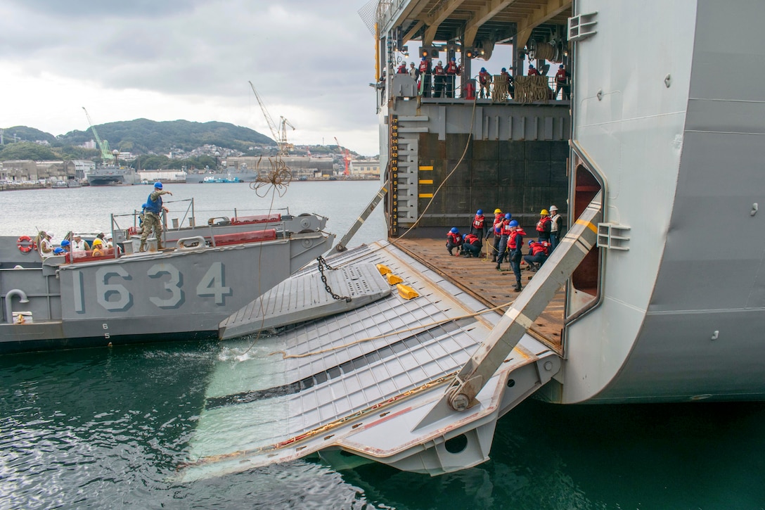 Sailors connect a landing craft to the open ramp of a ship in water.