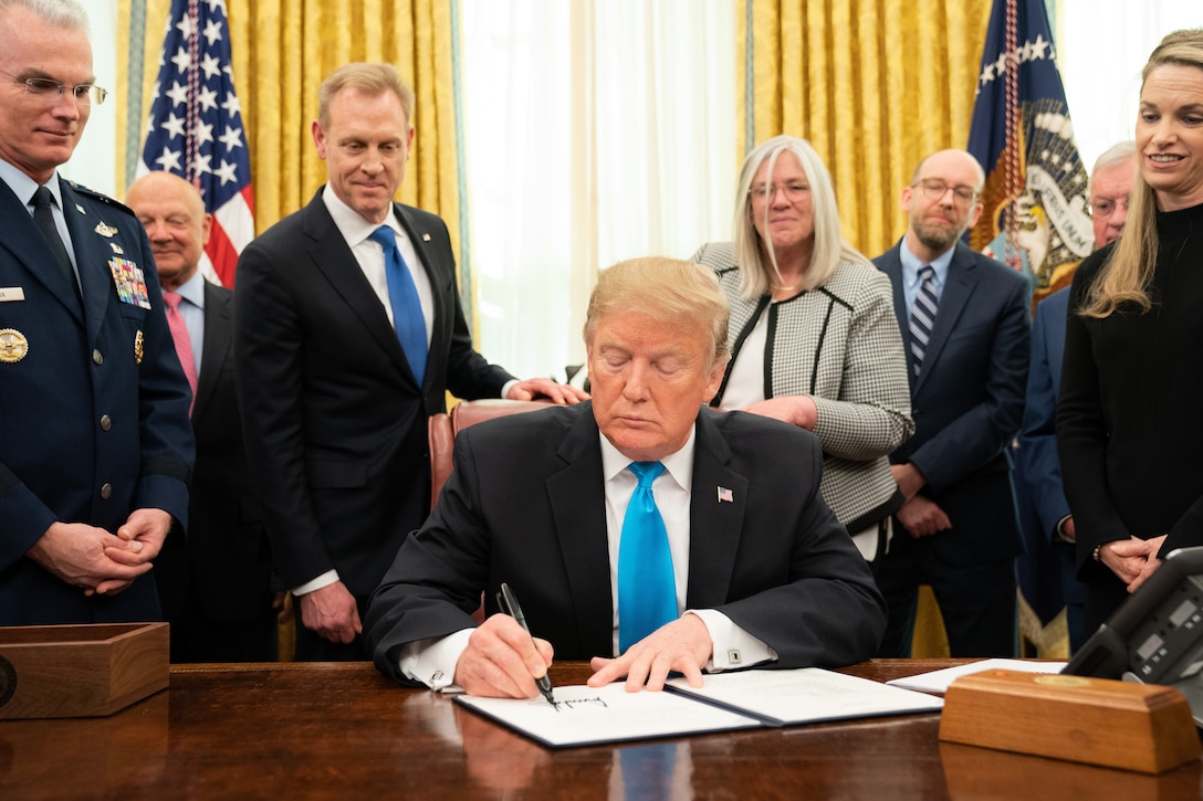 President Donald J. Trump signs a paper while sitting at his desk surrounded by people.