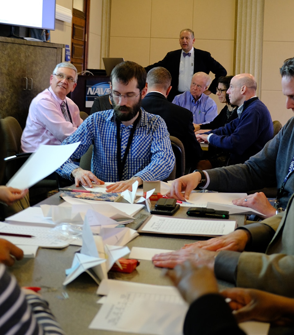 Dr. Nicholas Jones (center, front), a materials engineer in the Physical Metallurgy and Fire Performance Branch at Naval Surface Warfare Center, Carderock Division, works with his team making a paper airplane within specifications during a workshop for building systems on Feb. 14, 2019. The workshop was part of a visit by Dr. Steven Spear (standing), author of the best-selling book “The High-Velocity Edge,” to Carderock’s West Bethesda, Md., headquarters. (U.S. Navy photo by Kelley Stirling/Released)