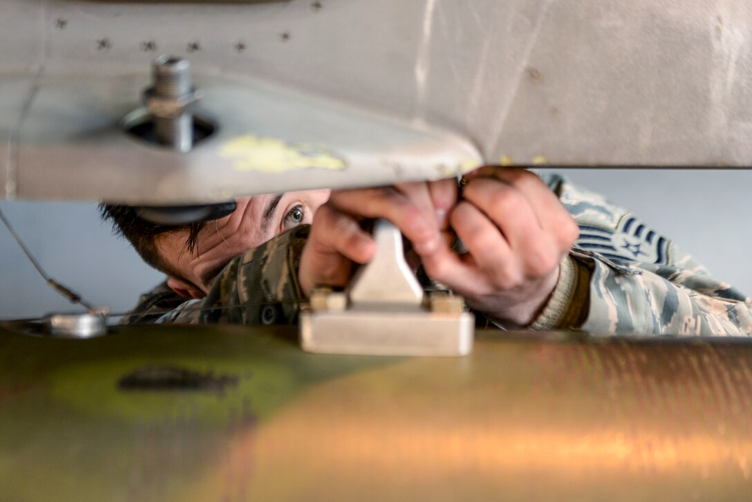 An airman's face is partially obscured between horizontal sections of metal and his hands as he does maintenance-type work.