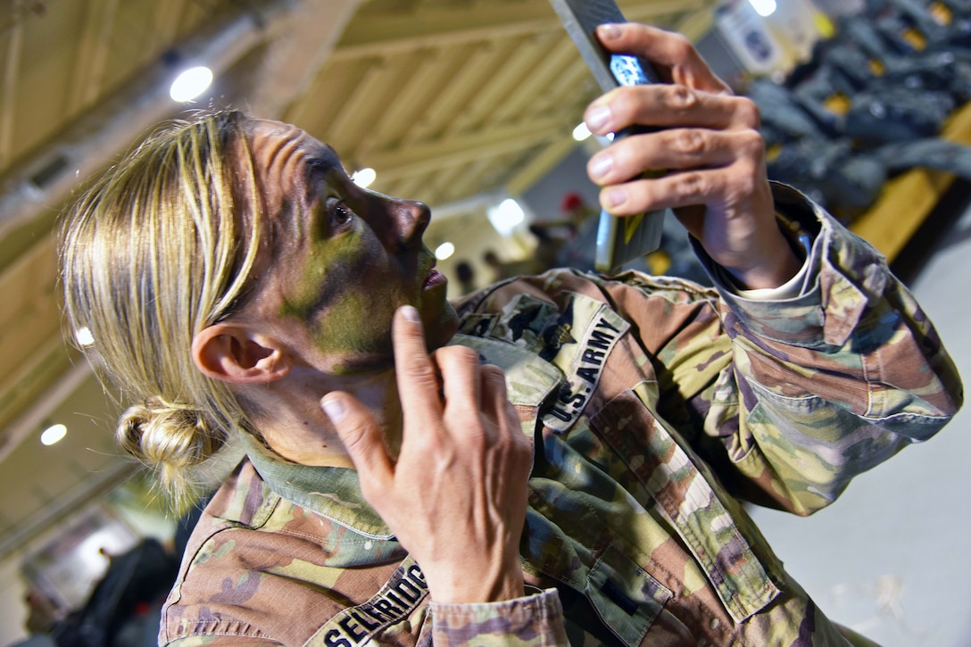 A soldier applies face paint while looking in a hand mirror.