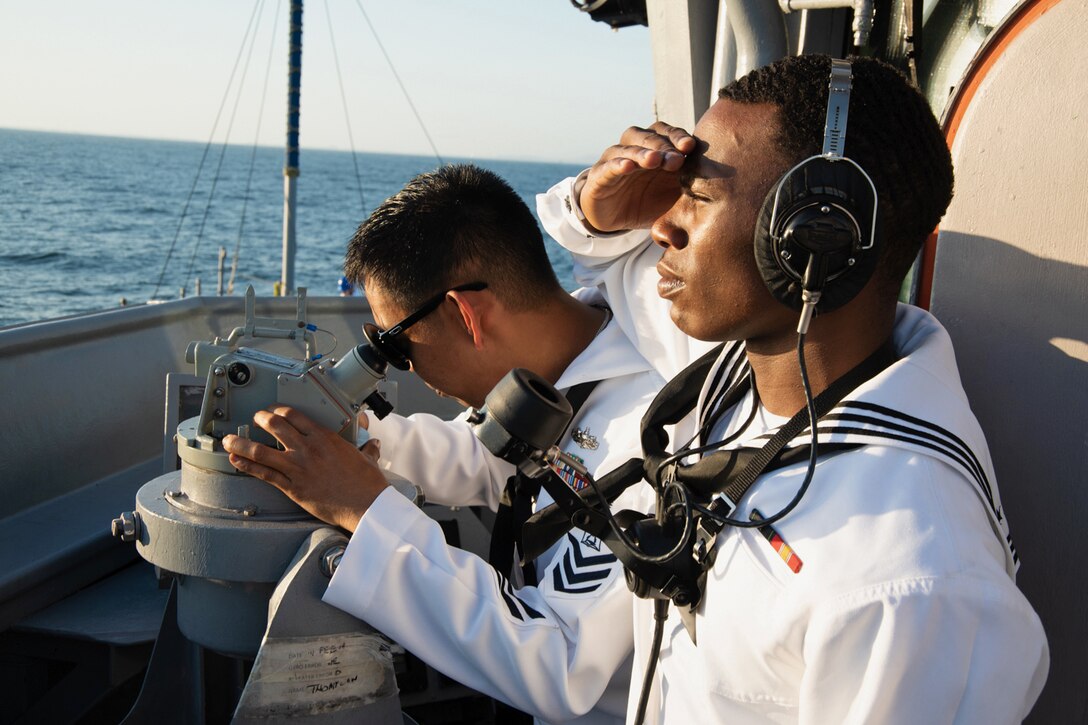 Two sailors look out to sea from the  deck of a ship.