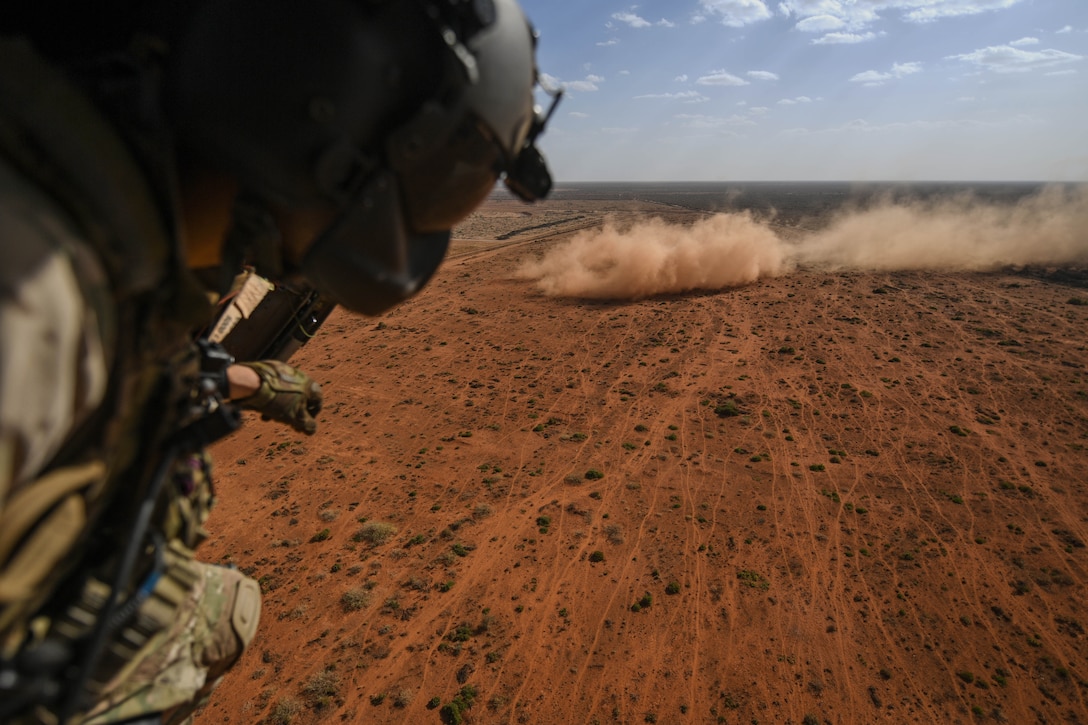 An airman in a helicopter looks out at dust and dry earth below.