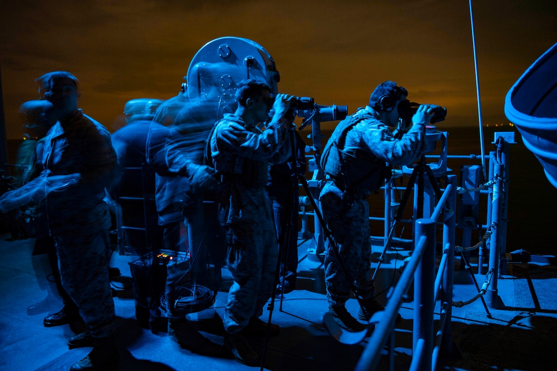 Service members, illuminated by blue light, stand watch on ship's deck, some with binoculars.