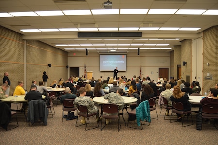 People sitting at round tables in the middle in big room. There is display screen in the background with Congressman Warren Davidson standing on a stage giving a speech. There are students and soldiers in uniform observing.