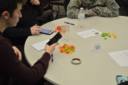 Students sitting at a round table counting gummy bears. One student is using his phone to count the gummy bears