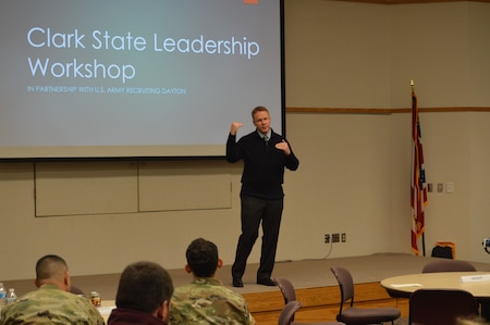 Congressman Warren Davidson speaking at the 2019 Leadership Conference and Workshop at Clark State Community College. He's wearing a black sweater vest with tie. There are soldiers sitting in front of him wearing multicam uniforms. There is a giant screen with the words 2019 Leadership Conference and Workshop behind the Congressman.