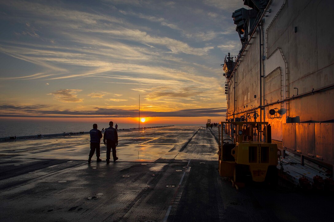 Sailors look at a sun setting beyond the ocean horizon.