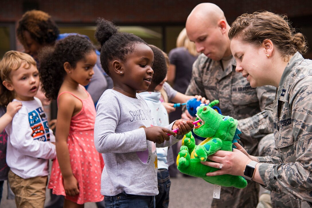 An airman holds a green stuffed animal while kneeling next to a child holding up a toothbrush.