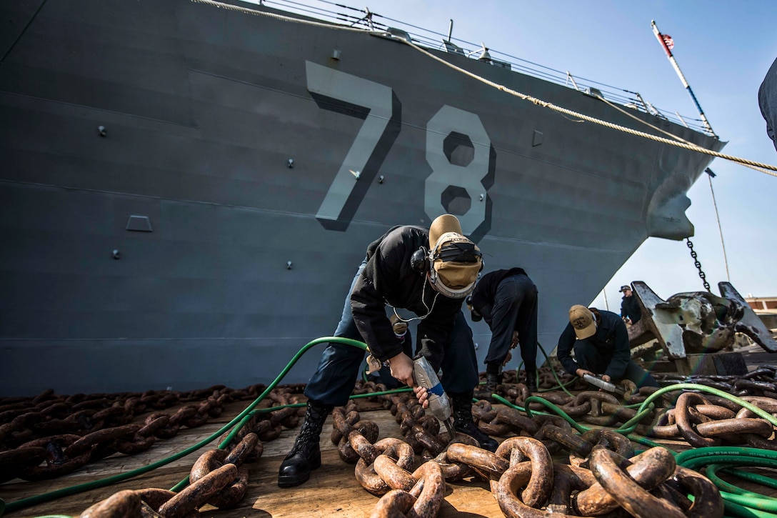 Sailors holding tools bend over to clean chain next to a ship.