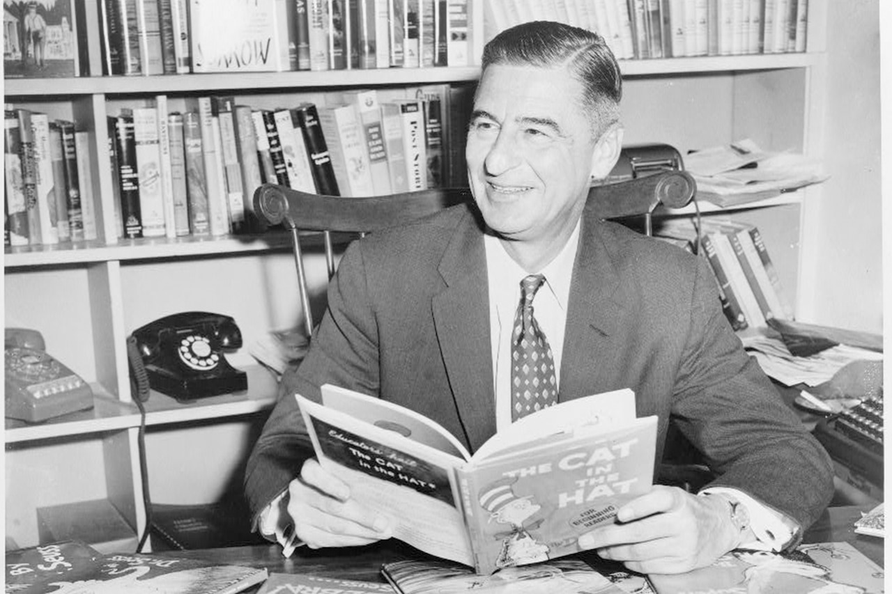 A man seated at desk covered with books holds a book and smiles.