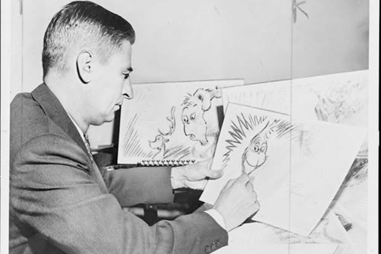 A man sits at a desk drawing cartoons on paper.
