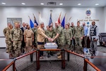 Military members and a few civilians stand in front of flags while the two senior members, civilian and military, cut a ceremonial cake.