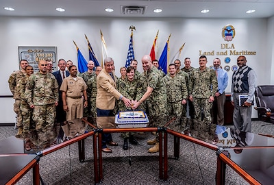 Military members and a few civilians stand in front of flags while the two senior members, civilian and military, cut a ceremonial cake.