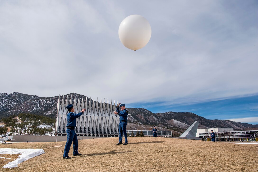 Two airmen hold onto a weather balloon in the sky with mountains in the background.