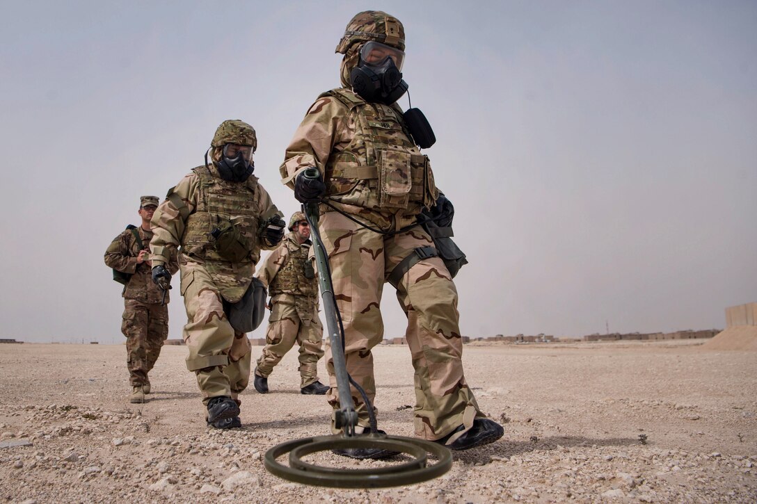 A soldier holds a device to the ground as three other people walk behind.