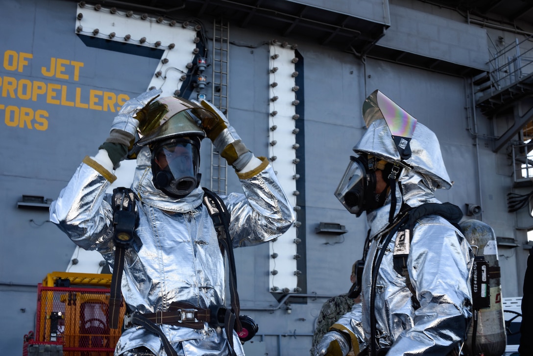 Two men wear crash salvage suits on the deck of a ship.