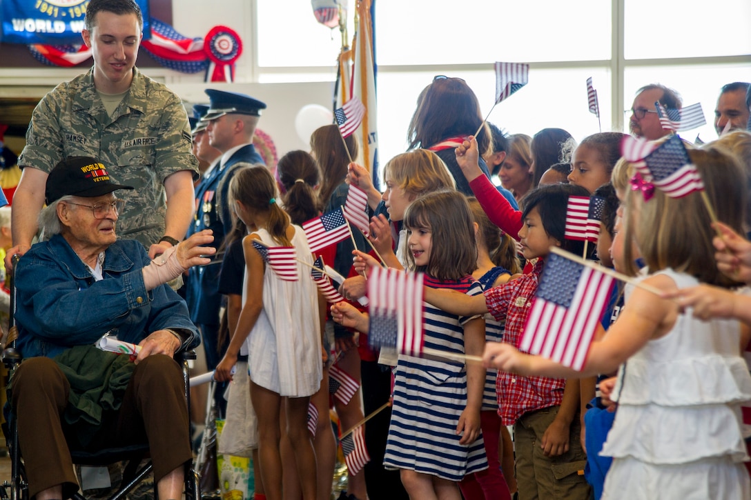 A group of children holding miniature American flags greet a veteran in a wheelchair being pushed by an airman.