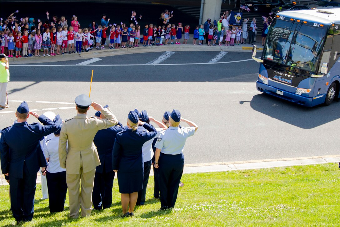 A group of people stand along  both sides of a road saluting and waving at a bus driving by.