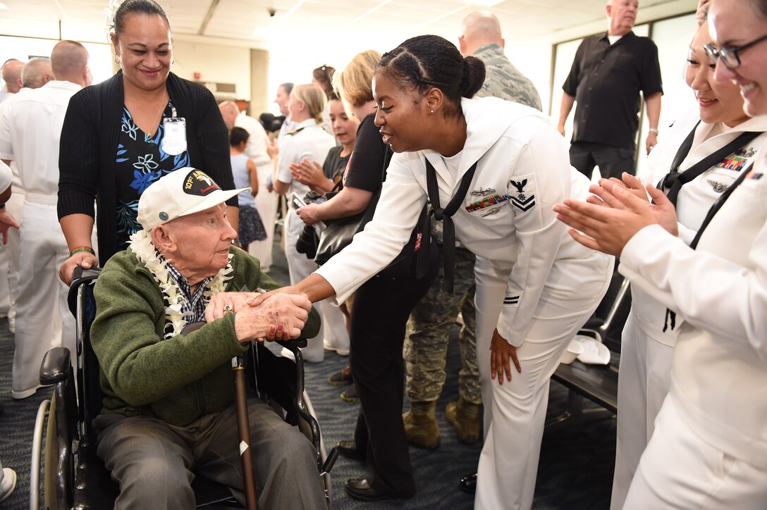 A man being pushed in a wheelchair by a woman shakes hands with sailors lined up in an airport terminal.