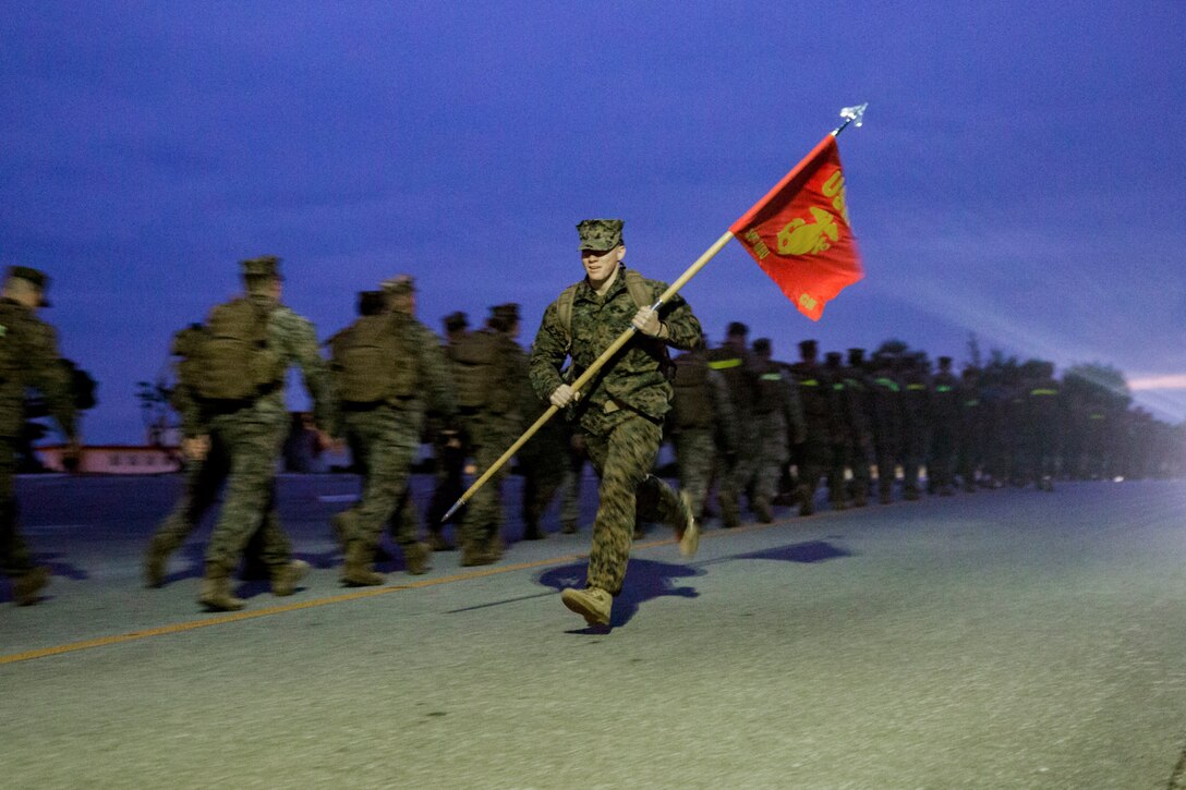 A Marine runs carrying a flag alone a line of other Marines.