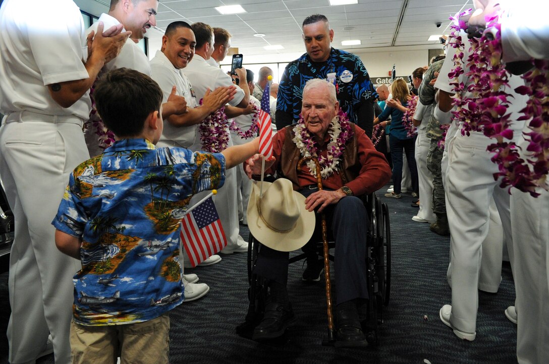 A child hands a man in a wheelchair an American flag as the man is being pushed in-between two rows of people.