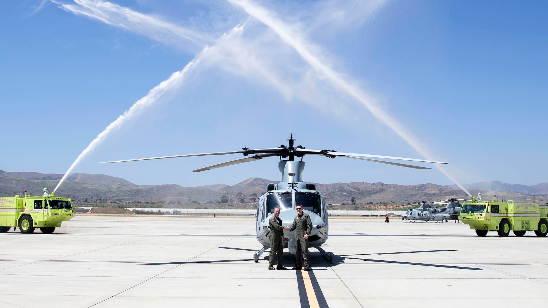 Two Marine pilots shake hands in front of the last UH-1y Venom delivery while two flight-line firetrucks spray their water cannons in the air.