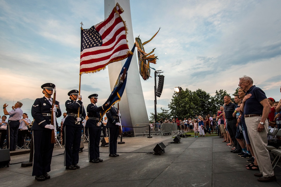 Two service members holding rifles and two service members holding flags stand in front of a group of people in a crowd holding their hands to their heart.